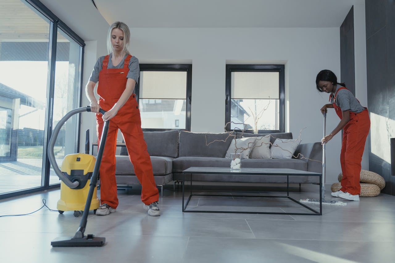 about-01 Two women in red overalls vacuuming and sweeping a modern living room.